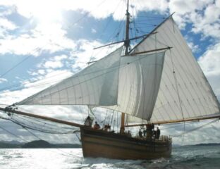 A replica of “The Restauration” Norwegian sailing ship shown at full sail on the sea with a bright blue sky with white clouds behind the sails.