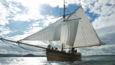 A replica of “The Restauration” Norwegian sailing ship shown at full sail on the sea with a bright blue sky with white clouds behind the sails.