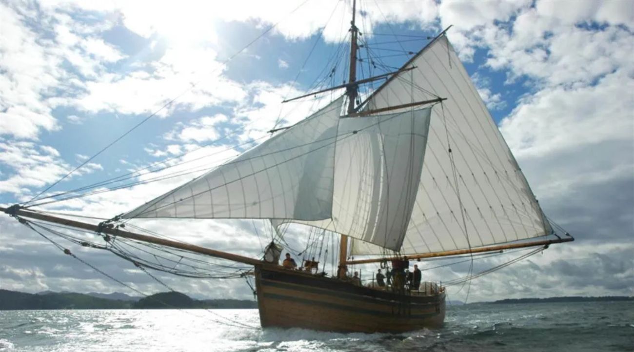 A replica of “The Restauration” Norwegian sailing ship shown at full sail on the sea with a bright blue sky with white clouds behind the sails.