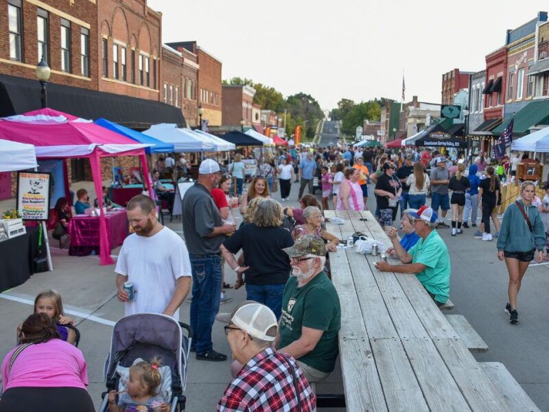 Small older main street with brightly colored tent set up as people gather, walk around, and visit with neighbors.