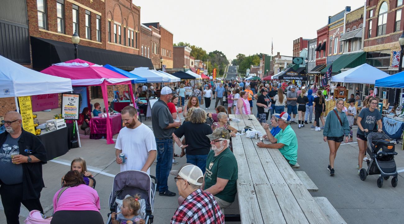 Small older main street with brightly colored tent set up as people gather, walk around, and visit with neighbors.