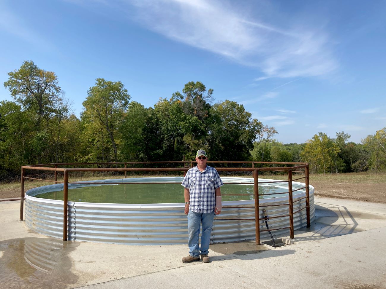 Dean Thomas dressed in a checked short-sleeve shirt stand next to a large 11,500-gallon grain bin sheet tank, which is the only water source for cattle grazing within the adjacent state wildlife management area.