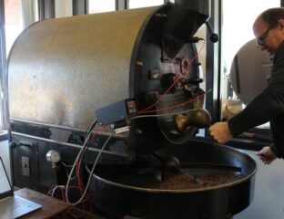 Middle aged man stand in front of a large black cast iron machine with a tray holding light brown coffee beans. A long arm moves through the beans as they roast.