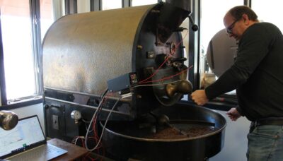 Middle aged man stand in front of a large black cast iron machine with a tray holding light brown coffee beans. A long arm moves through the beans as they roast.