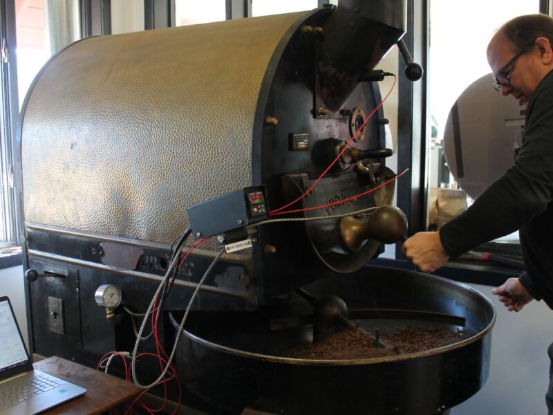 Middle aged man stand in front of a large black cast iron machine with a tray holding light brown coffee beans. A long arm moves through the beans as they roast.