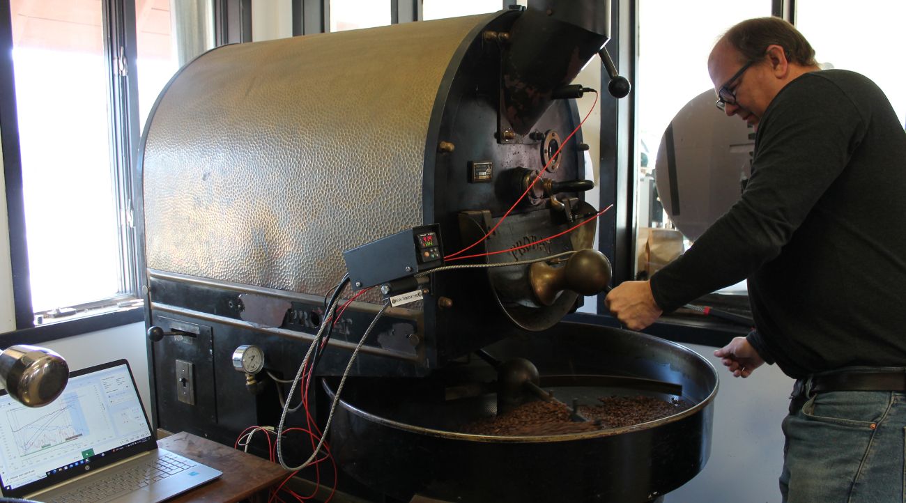 Middle aged man stand in front of a large black cast iron machine with a tray holding light brown coffee beans. A long arm moves through the beans as they roast.