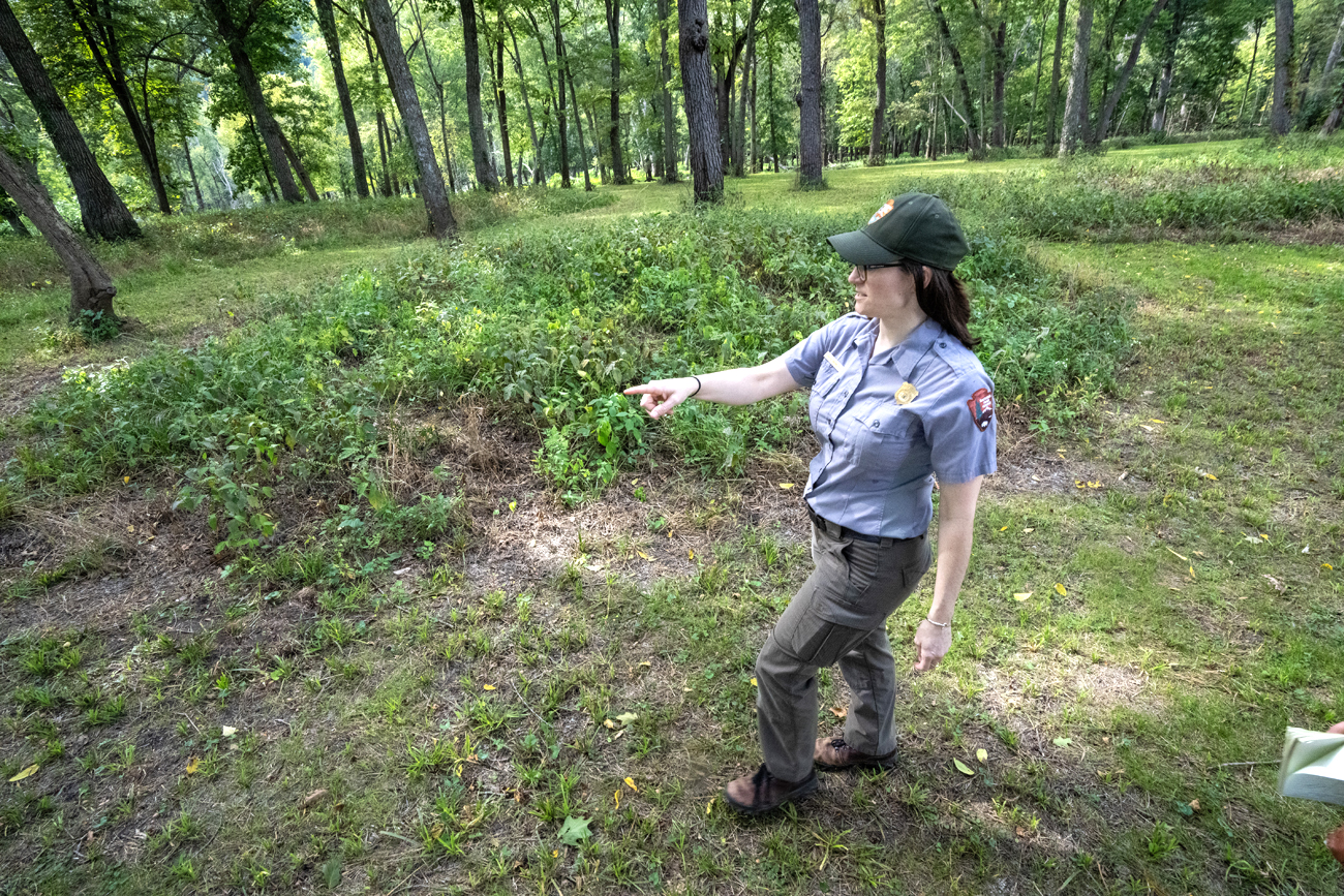 Woman in park officer uniform points to the ground that is slightly higher than surrounding area and covered in vegetation.