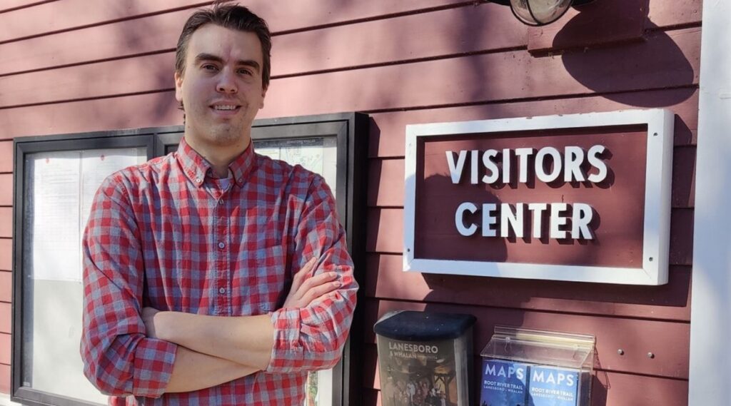 Man with red checked shirt stands with arms folded in front of a red wooden building by a sign that says Visitors Center.