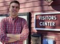 Man with red checked shirt stands with arms folded in front of a red wooden building by a sign that says Visitors Center.