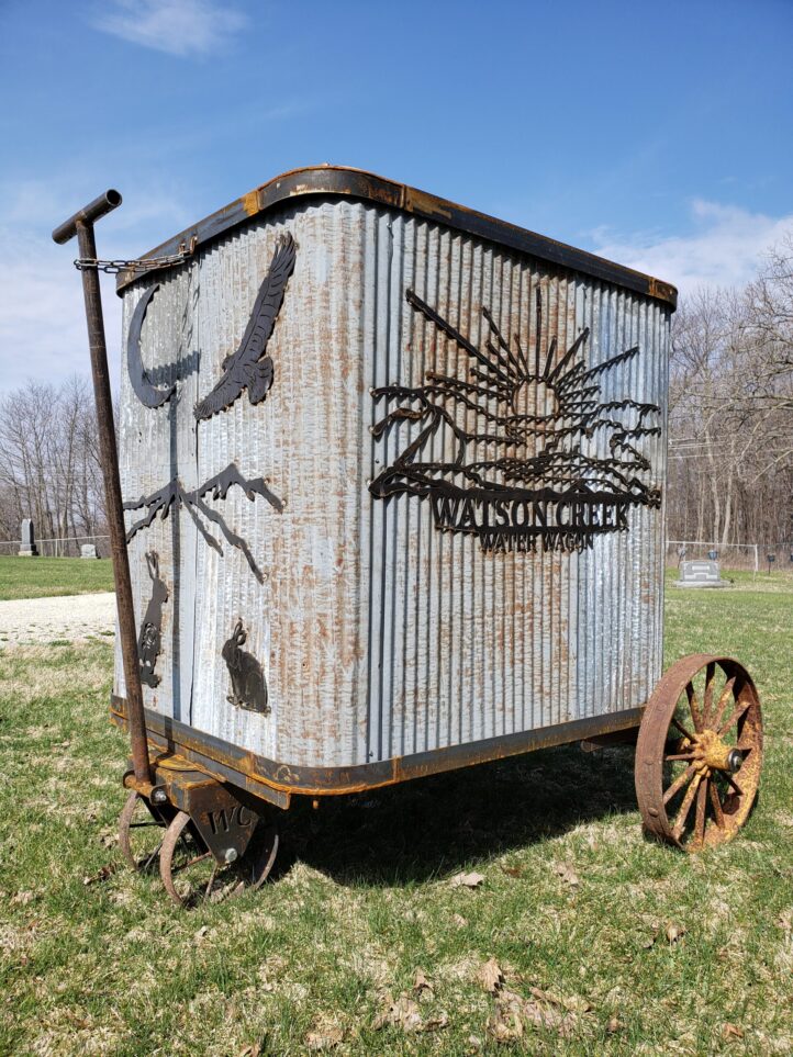 A large corrugated metal box with rusty metal frame, long handle, and wheels. Box has black and rusted metal art attached to the outside of animals, birds and a sunrise.