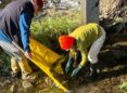 Two people dressed in jackets, hats, and rubber boots stand in shallow water and scoop debris from the bottom of the stream onto a yellow tarp.