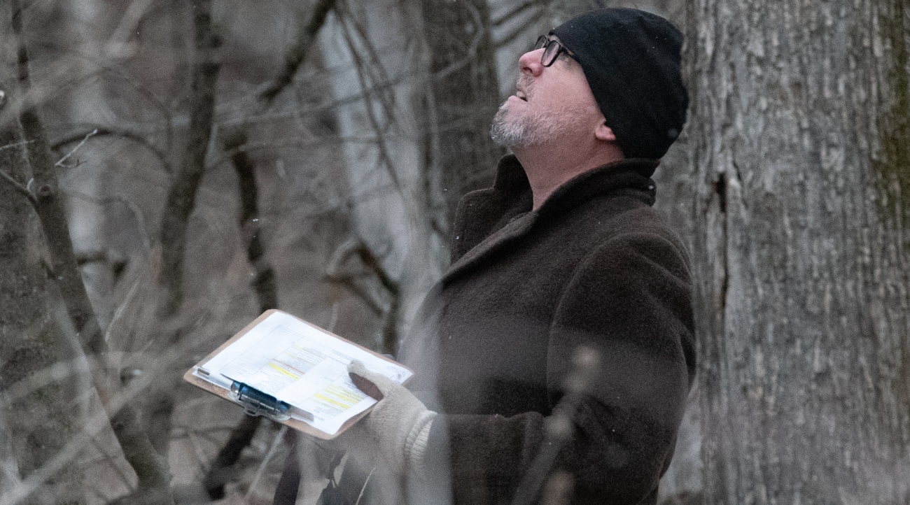 Man in thick overcoat with stocking cap and glasses looks upward while standing in a wooded area and holding a clipboard with a bird count paper.