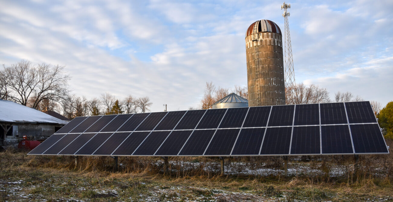 An array of solar panels in front of an old cement block farm silo against a blue sky with white and gray clouds.