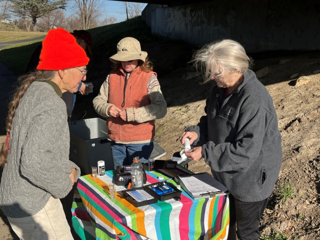 Three older women stand around a folding table next to the trout stream. They are looking at water testing supplies and getting them set up for their work.