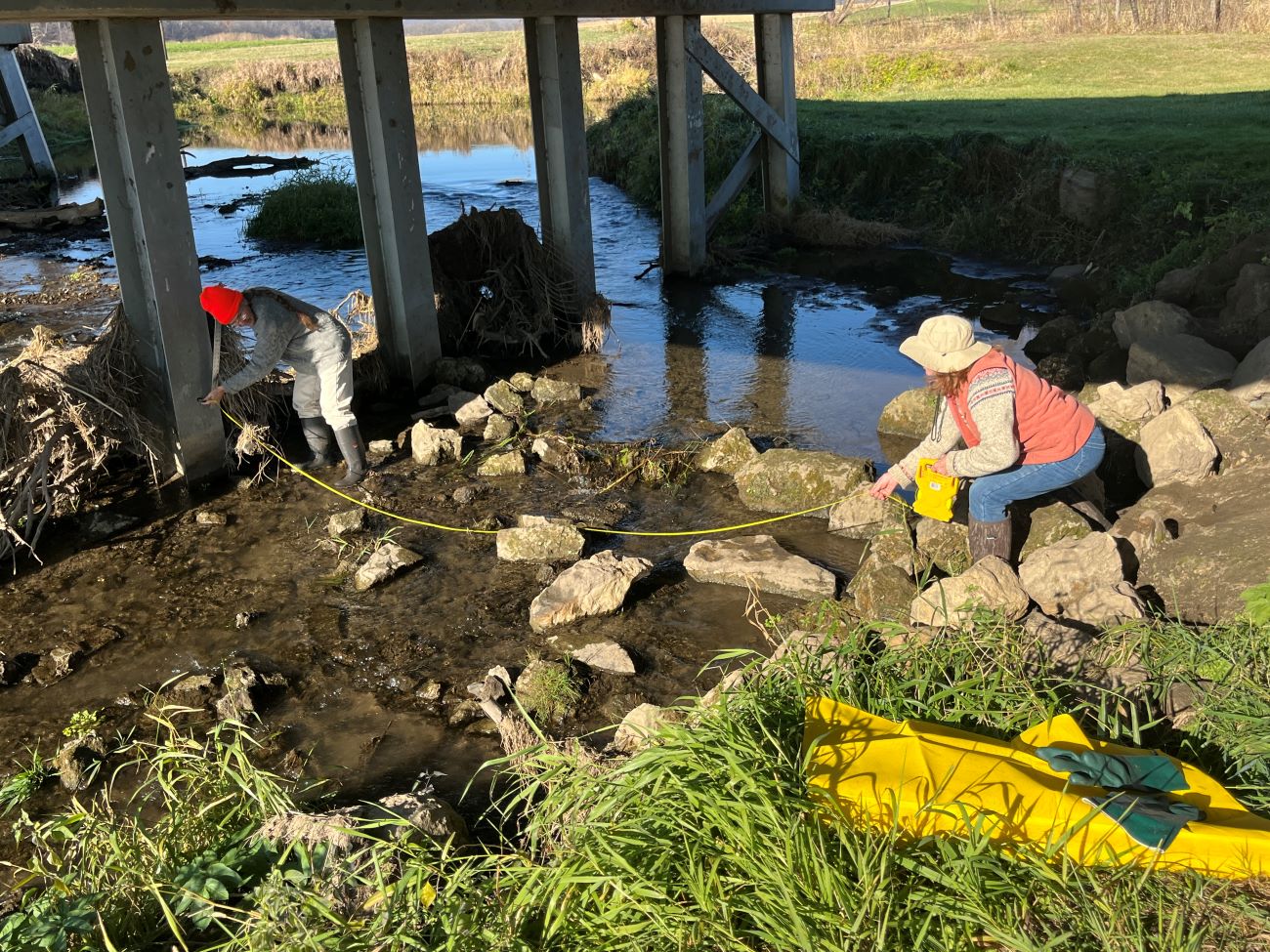 Two adults dressed in outdoor gear stand on either side of shallow stream. They are holding a measuring tape to measure the width of the stream.