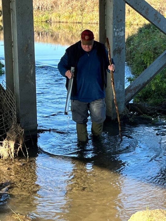 A man dressed in winter clothing and rubber boots wades in a shallow water holding a tube and testing equipment for water turbidity.