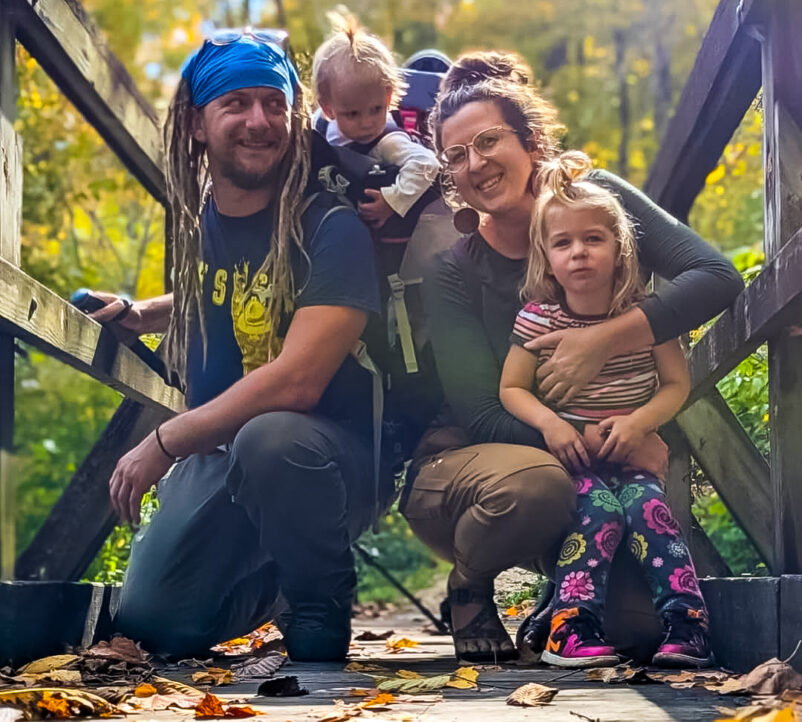 A family of a male wearing a blue bandana with dreadlocks, two young children, woman with gray and green top and pants crouch closely together against a wooden bridge frame.