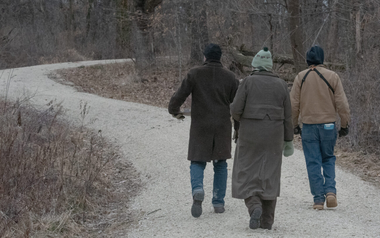 A group of three people dressed in winter clothes walk away from the camera along a winding gravel road through a wooded area.