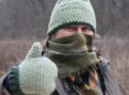 A woman bundled up with a knitted green hat, knitted green mittens, and a plaid scarf gives a thumbs up outside on a winter day.