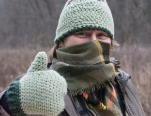 A woman bundled up with a knitted green hat, knitted green mittens, and a plaid scarf gives a thumbs up outside on a winter day.