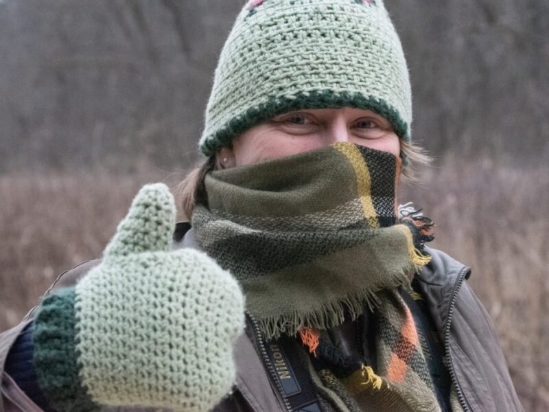 A woman bundled up with a knitted green hat, knitted green mittens, and a plaid scarf gives a thumbs up outside on a winter day.