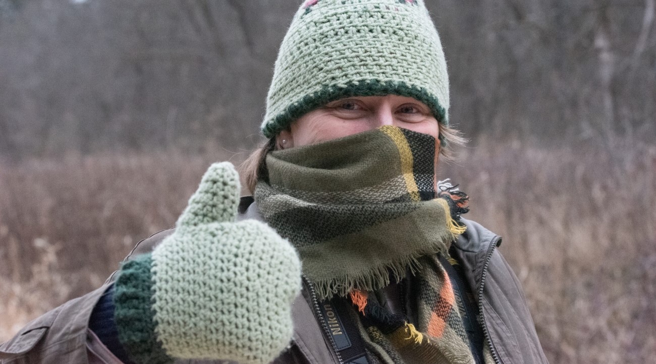 A woman bundled up with a knitted green hat, knitted green mittens, and a plaid scarf gives a thumbs up outside on a winter day.