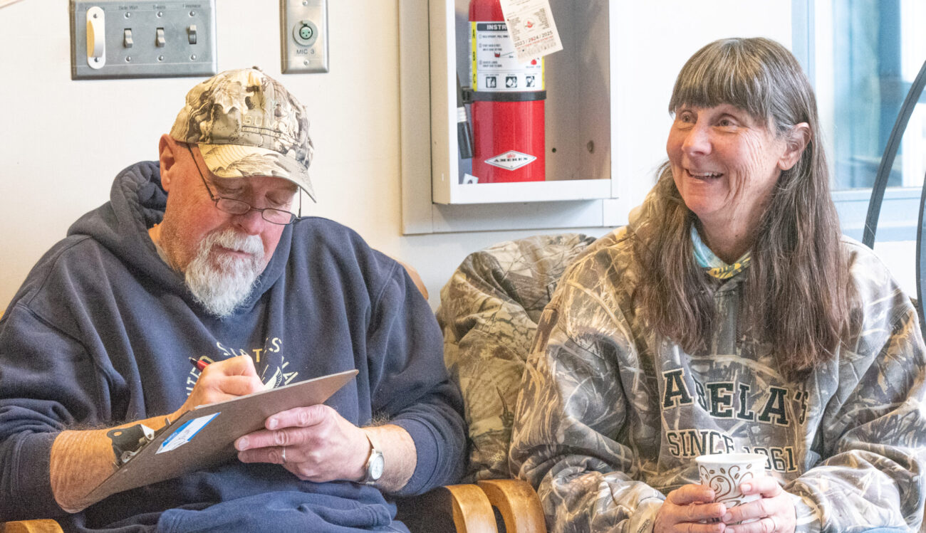 An older man writes on a piece of paper while his wife with long brown hair looks to the side.