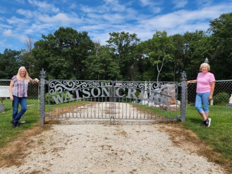 Two women stand in front of an iron gate with the words Watson Creek Cemetery