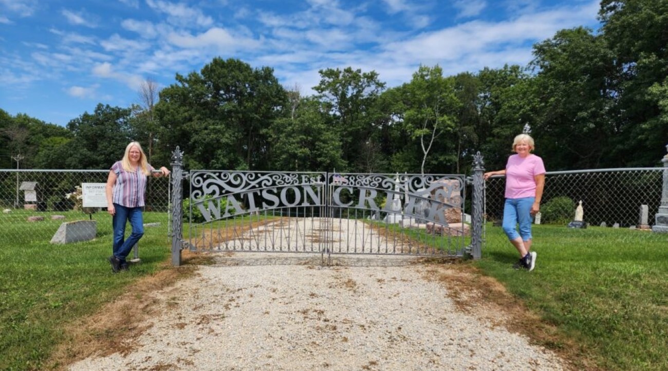 Two women stand in front of an iron gate with the words Watson Creek Cemetery