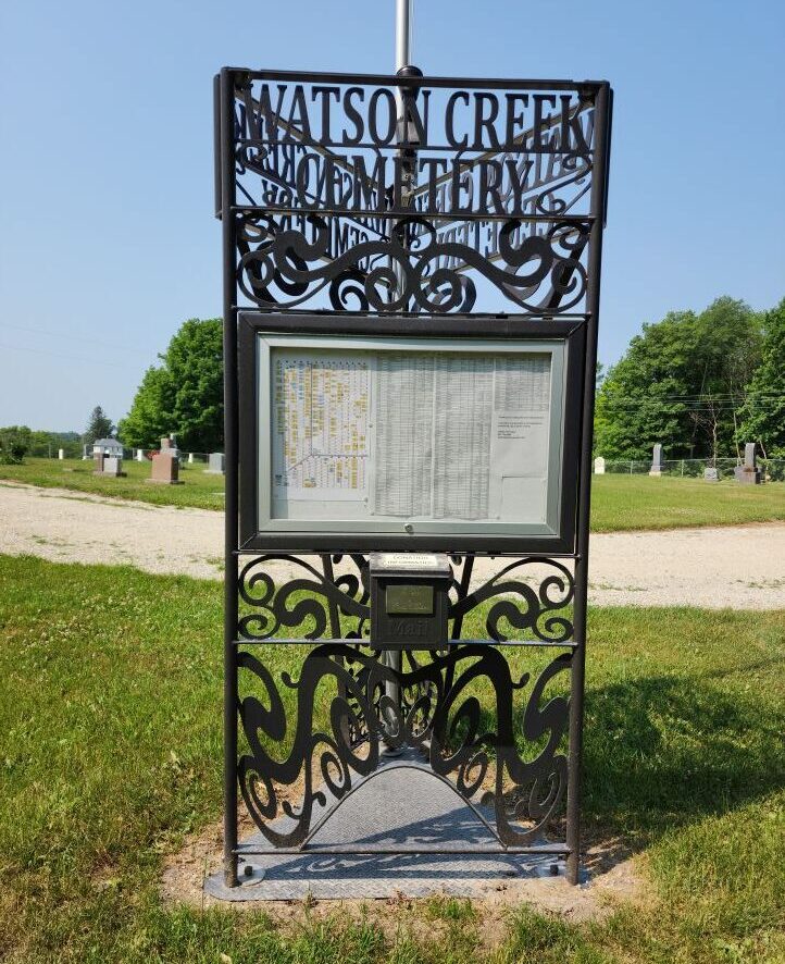 A tall, narrow black metal frame with metal scroll work and the words Watson Creem Cemetery. In the middle is a frame and plexiglass covering the map of the cemetery.