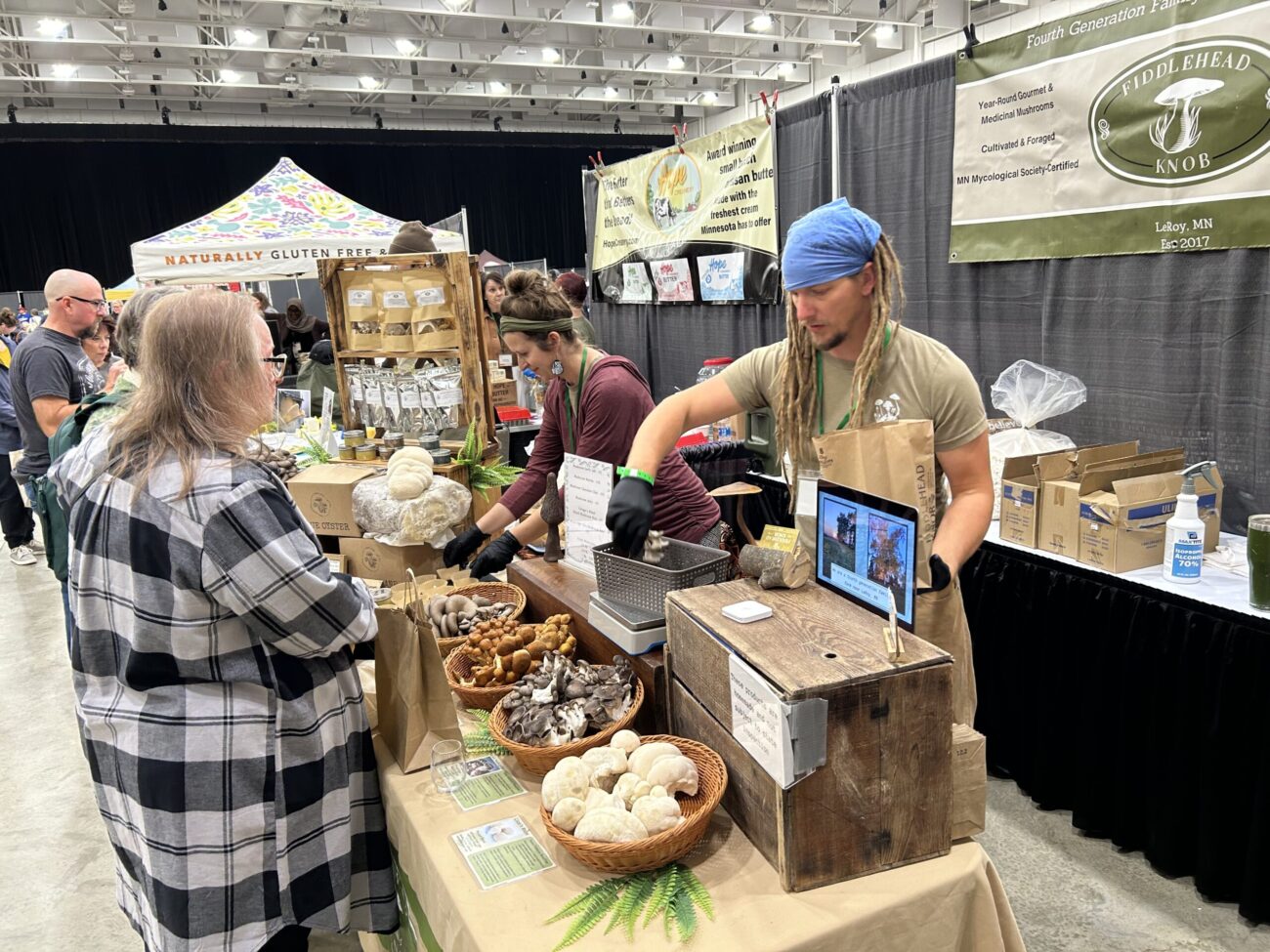 A man and woman stand behind their table of mushroom products and speak with a customer, a woman dressed in a flannel shirt.