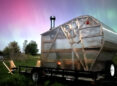 A bright pink, purple, blue, and green sky of Northern Lights shine behind a plexiglas and wood sauna.