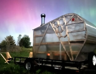 A bright pink, purple, blue, and green sky of Northern Lights shine behind a plexiglas and wood sauna.