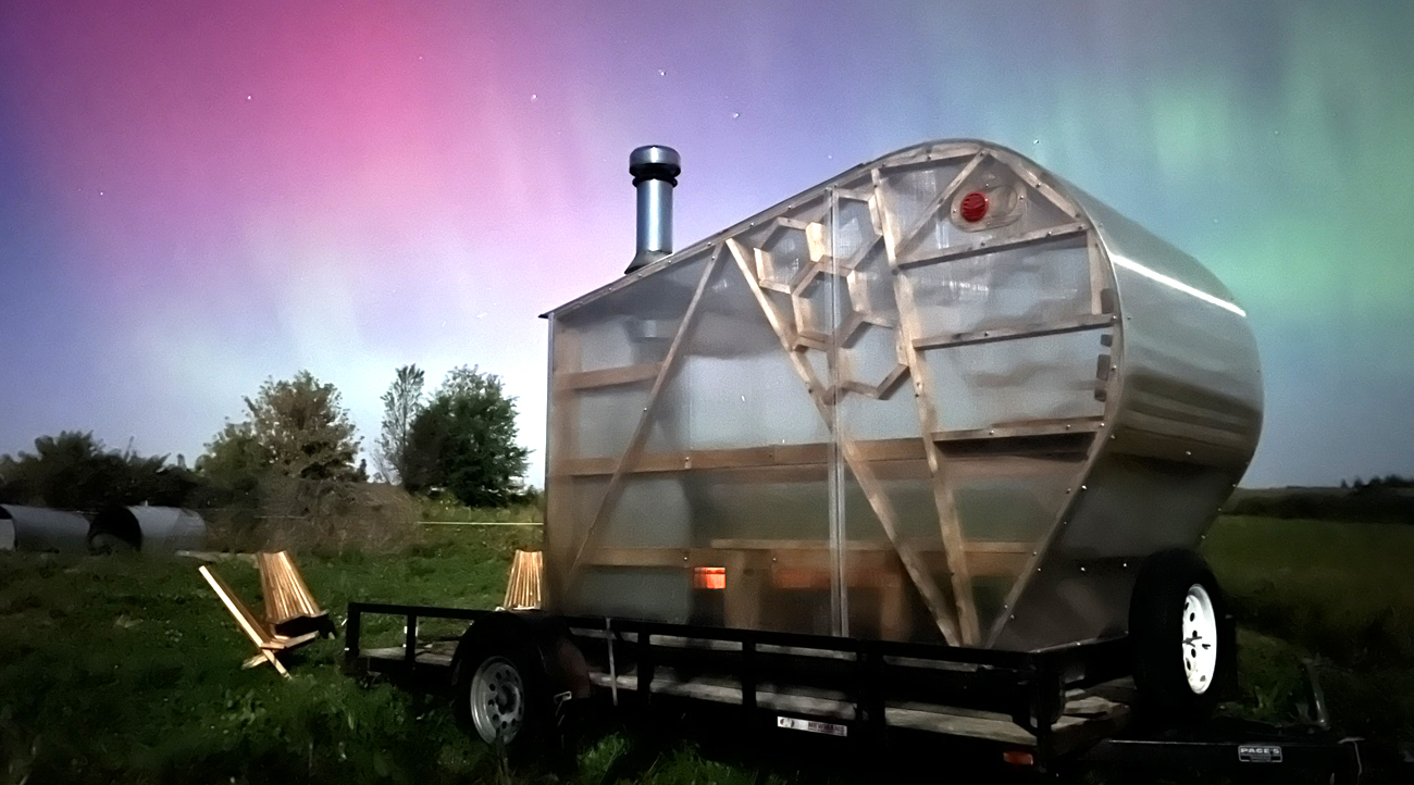 A bright pink, purple, blue, and green sky of Northern Lights shine behind a plexiglas and wood sauna.