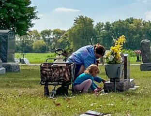 A woman and a small child kneel next to a gravestone. Beside them is a wagon holding cleaning supplies. Grass is green, sky is blue.