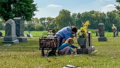 A woman and a small child kneel next to a gravestone. Beside them is a wagon holding cleaning supplies. Grass is green, sky is blue.