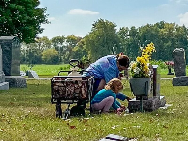 A woman and a small child kneel next to a gravestone. Beside them is a wagon holding cleaning supplies. Grass is green, sky is blue.