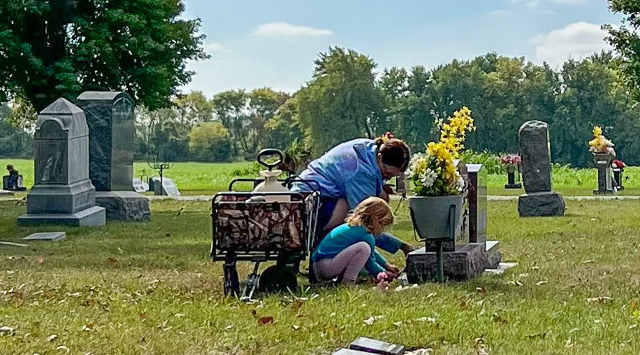 A woman and a small child kneel next to a gravestone. Beside them is a wagon holding cleaning supplies. Grass is green, sky is blue.