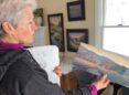 A woman with short gray hair holds a small rectangular, unframed landscape painting in front of her.