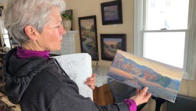 A woman with short gray hair holds a small rectangular, unframed landscape painting in front of her.