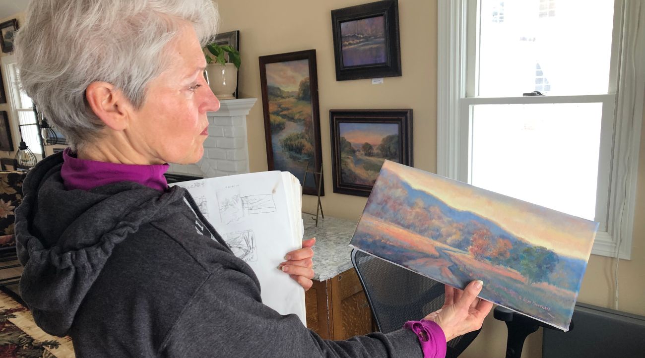 A woman with short gray hair holds a small rectangular, unframed landscape painting in front of her.