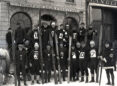 Black and white photo of a group of downhill skiers holding their skis upright in front of them with dark uniforms and large white labels on the front with their team numbers.