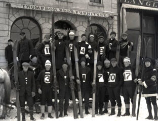 Black and white photo of a group of downhill skiers holding their skis upright in front of them with dark uniforms and large white labels on the front with their team numbers.