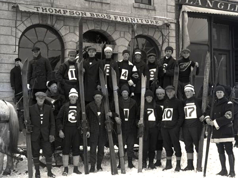 Black and white photo of a group of downhill skiers holding their skis upright in front of them with dark uniforms and large white labels on the front with their team numbers.