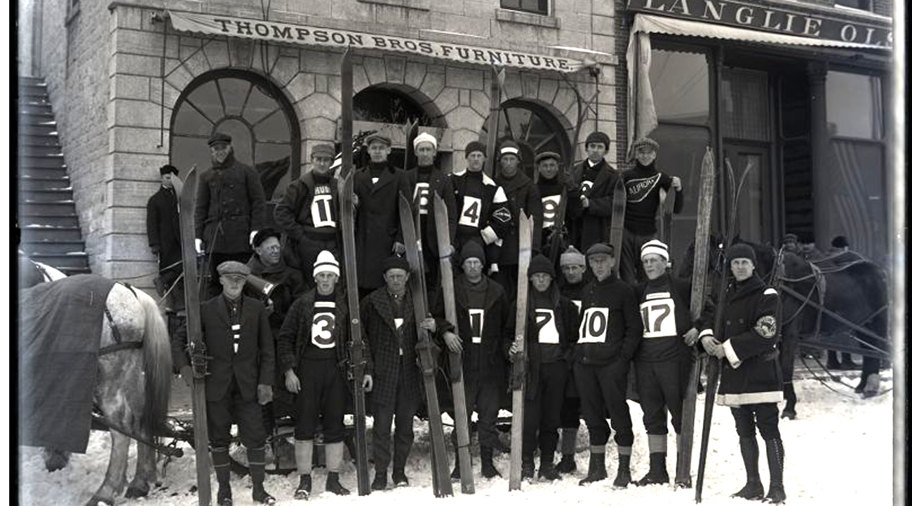 Black and white photo of a group of downhill skiers holding their skis upright in front of them with dark uniforms and large white labels on the front with their team numbers.