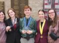 Two adult women stand on either side of three high school students wearing medals around their necks. The boy in the middle is holding a crystal plaque.