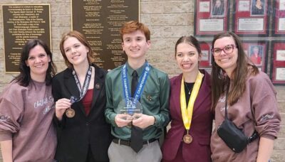 Two adult women stand on either side of three high school students wearing medals around their necks. The boy in the middle is holding a crystal plaque.