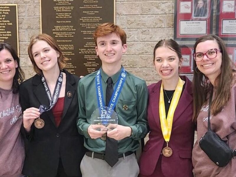 Two adult women stand on either side of three high school students wearing medals around their necks. The boy in the middle is holding a crystal plaque.