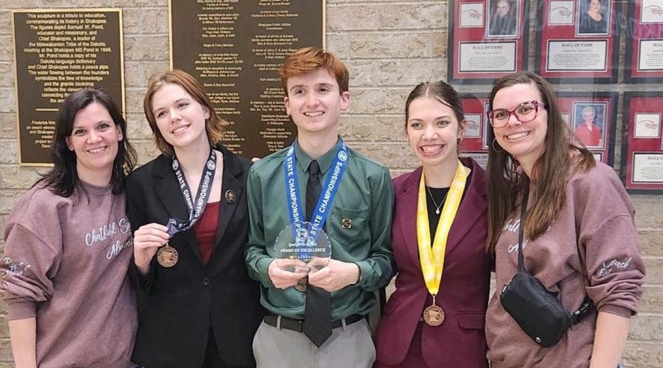 Two adult women stand on either side of three high school students wearing medals around their necks. The boy in the middle is holding a crystal plaque.