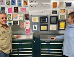 A man in a brown shirt and woman in a light blue sweater stand in front of dark green cabinets and small pieces of art.